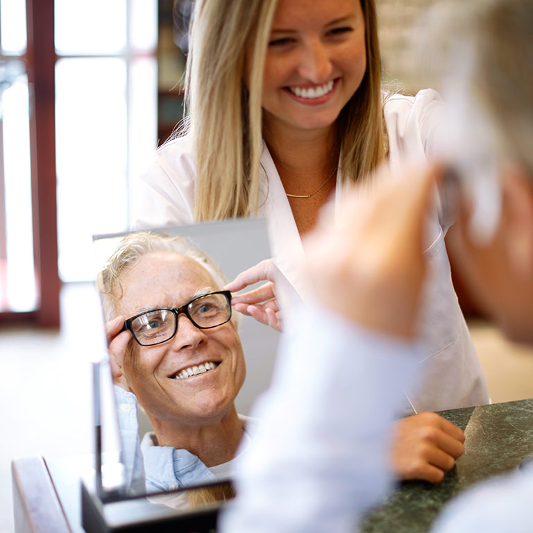 A male customer tries on a pair of black rimmed glasses at the Atwater Eye Care Center. He is looking in the mirror while being helped by a female staff member.