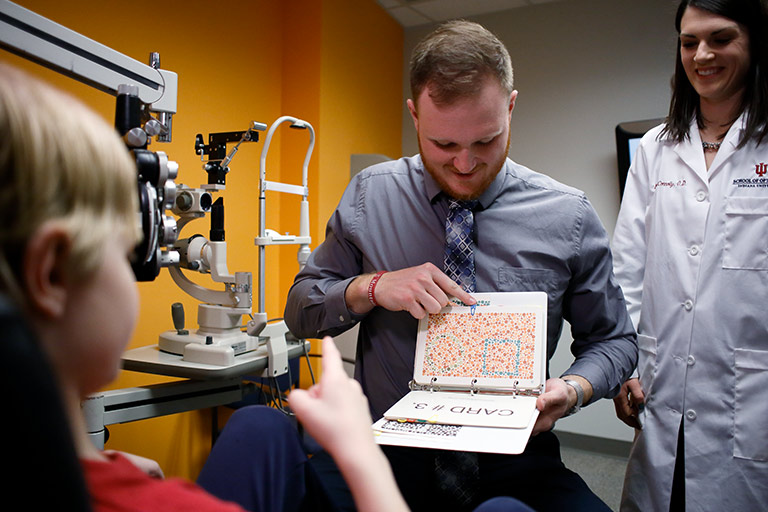 A male optometry student in shirt and tie holds up a binder of colored dot designs. The patient, a six-year-old boy, points to the page.