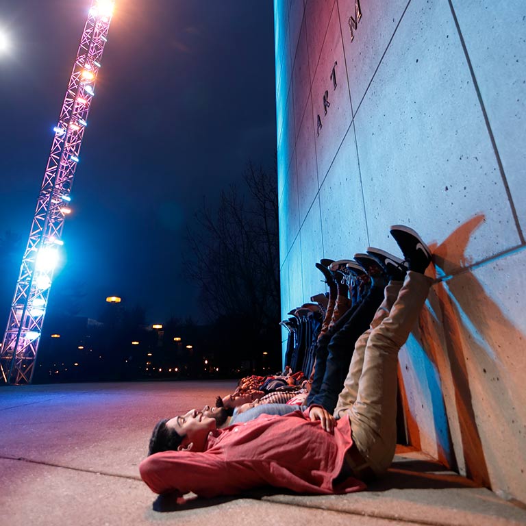 A diverse group of students lay on the ground in front of the art museum, lit by the famous Light Totem.